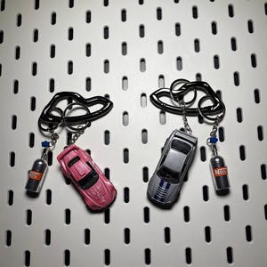 Peut inclure: Deux porte-clés de voiture avec des anneaux noirs. L'un a une voiture rose et une bouteille NOS argentée. L'autre a une voiture argentée avec des rayures bleues et une bouteille NOS argentée. Les porte-clés sont suspendus à un panneau perforé blanc.
