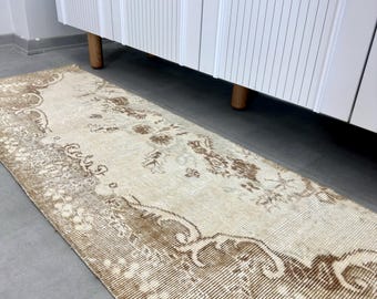 May include: A rectangular, distressed-look rug with a beige and brown floral pattern. The rug has a border with a detailed design and is placed on a gray floor. The rug is positioned in front of a white cabinet with wooden legs.