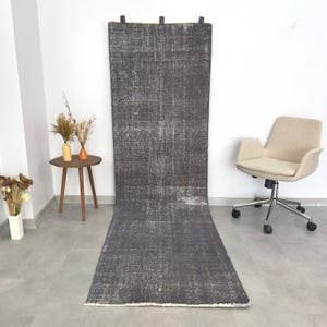 Op de afbeelding: Een lange, rechthoekige, verweerde grijze vloerkleed met witte accenten. Het vloerkleed hangt aan een witte muur met zwarte bevestigingen aan de bovenkant. Het vloerkleed ligt op een grijze vloer. Een kleine houten tafel met vazen en een beige bureaustoel staan op de achtergrond.