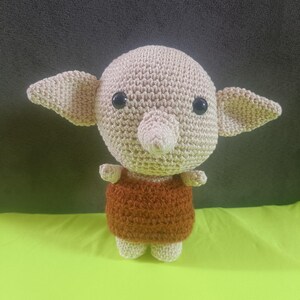 May include: A handmade crochet doll with a round head, large ears, and a small nose. The doll has black eyes and is wearing a brown outfit. The doll is sitting on a green surface.