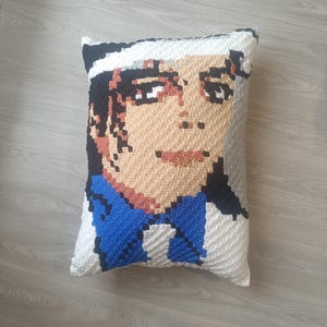 Peut inclure: Un coussin crocheté à la main représentant un portrait pixélisé d'une personne à la peau bronzée, aux cheveux noirs et portant un chapeau blanc. Le coussin a une chemise bleue et blanche. Le coussin est rectangulaire.