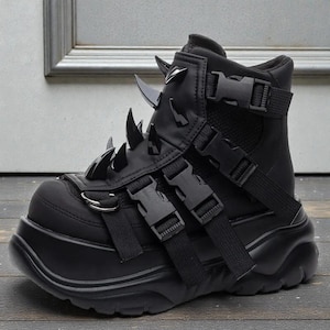 May include: Black platform boot with multiple straps, buckles, and decorative spikes. The high-top boot features a thick sole and a combination of textures, including smooth and mesh materials. The boot has a futuristic, edgy design.