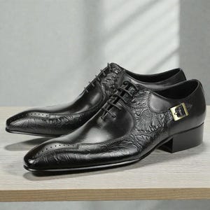 Pu&ograve; includere: Un paio di scarpe eleganti in pelle nera con dettagli floreali in rilievo. Le scarpe presentano un classico design stringato, una punta affusolata e una piccola fibbia color oro sul lato. Le scarpe sono esposte su una superficie di legno.