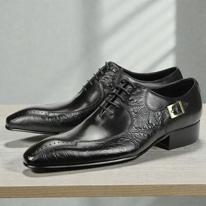 Pu&ograve; includere: Un paio di scarpe eleganti in pelle nera con dettagli floreali in rilievo. Le scarpe presentano un classico design stringato, una punta affusolata e una piccola fibbia color oro sul lato. Le scarpe sono esposte su una superficie di legno.