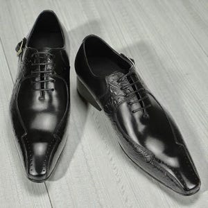 Pu&ograve; includere: Un paio di scarpe eleganti in pelle nera con finitura lucida. Le scarpe presentano una punta affusolata, chiusura con lacci e dettagli decorativi in rilievo sulla tomaia e sui lati. Un dettaglio con fibbia &egrave; visibile sul lato di una scarpa.
