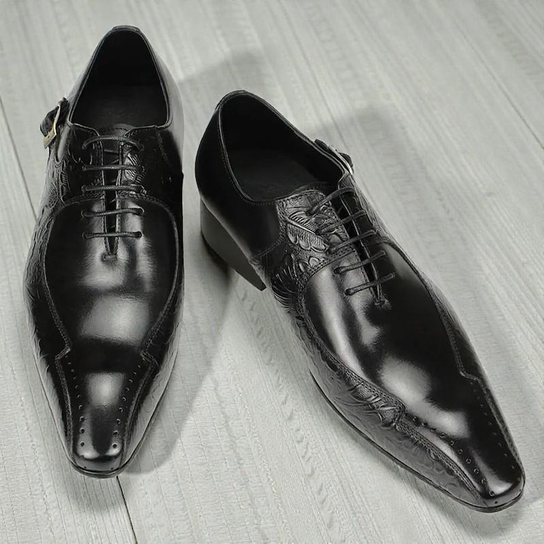 Pu&ograve; includere: Un paio di scarpe eleganti in pelle nera con finitura lucida. Le scarpe presentano una punta affusolata, chiusura con lacci e dettagli decorativi in rilievo sulla tomaia e sui lati. Un dettaglio con fibbia &egrave; visibile sul lato di una scarpa.
