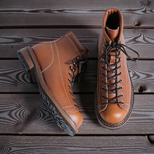 May include: Two brown leather boots with black laces are displayed on a wooden surface. The boots have a classic design with a rounded toe, a sturdy sole, and a lace-up closure. The leather has a textured appearance.