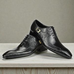 Pu&ograve; includere: Un paio di scarpe eleganti in pelle nera con dettagli floreali in rilievo. Le scarpe presentano una punta affusolata, chiusura con lacci e fibbia color oro. Le scarpe sono esposte su una superficie di legno chiaro.