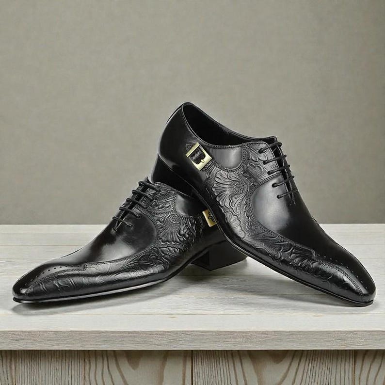 Pu&ograve; includere: Un paio di scarpe eleganti in pelle nera con dettagli floreali in rilievo. Le scarpe presentano una punta affusolata, chiusura con lacci e fibbia color oro. Le scarpe sono esposte su una superficie di legno chiaro.