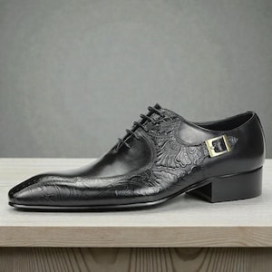 Pu&ograve; includere: Scarpa da uomo in pelle nera con dettagli floreali in rilievo. Presenta una punta affilata, chiusura con lacci e fibbia dorata. La scarpa ha un piccolo tacco a blocco ed &egrave; appoggiata su una superficie in legno.
