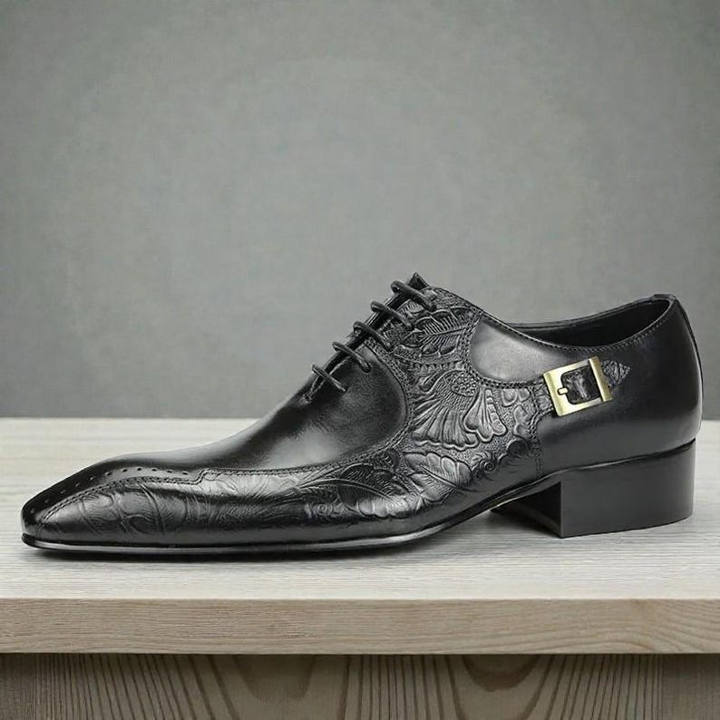 Pu&ograve; includere: Scarpa da uomo in pelle nera con dettagli floreali in rilievo. Presenta una punta affilata, chiusura con lacci e fibbia dorata. La scarpa ha un piccolo tacco a blocco ed &egrave; appoggiata su una superficie in legno.