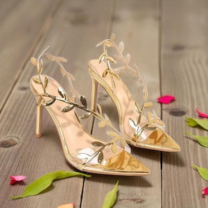 May include: A pair of gold high-heeled shoes with a pointed toe. The shoes feature a leaf design on the straps and a clear panel. The heels are approximately 4 inches tall. The shoes are on a wooden surface.