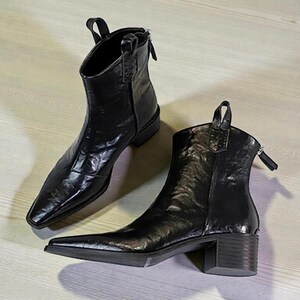 以下が含まれることがあります： 黒い革のアンクルブーツのペア。つま先は尖っており、小さなブロックヒールが付いています。ブーツには背面にプルタブとジッパー開閉部があります。光沢のある仕上げと洗練されたデザインが、モダンでスタイリッシュな外観を与えています。