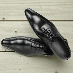 Pu&ograve; includere: Un paio di scarpe eleganti in pelle nera con finitura lucida. Le scarpe presentano un design stringato, perforazioni decorative e un motivo strutturato sui lati. Le scarpe sono appoggiate su una superficie di legno chiaro.