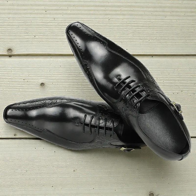 Pu&ograve; includere: Un paio di scarpe eleganti in pelle nera con finitura lucida. Le scarpe presentano un design stringato, perforazioni decorative e un motivo strutturato sui lati. Le scarpe sono appoggiate su una superficie di legno chiaro.