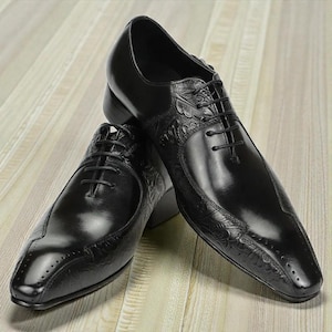 Pu&ograve; includere: Un paio di scarpe eleganti in pelle nera con dettagli in rilievo. Le scarpe presentano un design classico con lacci e una finitura lucida. Sono esposte su una superficie di legno chiaro.