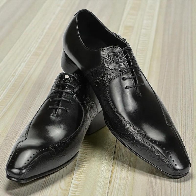 Pu&ograve; includere: Un paio di scarpe eleganti in pelle nera con dettagli in rilievo. Le scarpe presentano un design classico con lacci e una finitura lucida. Sono esposte su una superficie di legno chiaro.