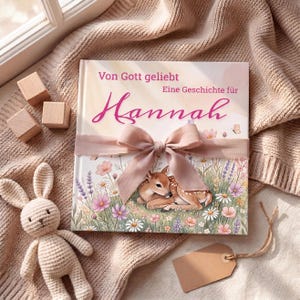 K&ouml;nnte beinhalten: Ein Kinderbuch mit dem Titel "Von Gott geliebt Eine Geschichte f&uuml;r Hannah" mit rosa Schleife und Schleife. Das Cover zeigt eine Rehhirsch-Illustration in einem Blumenfeld. Ein geh&auml;keltes Kaninchen und Holzbl&ouml;cke liegen neben dem Buch.