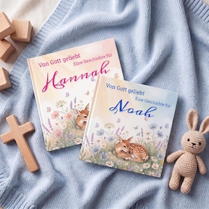 K&ouml;nnte beinhalten: Zwei Kinderb&uuml;cher mit den Namen "Hannah" und "Noah" auf den Einb&auml;nden, mit Illustrationen eines Rehs in einem Blumenfeld. Die B&uuml;cher liegen auf einer blauen Decke, zusammen mit einem Holzkreuz und einem Stoffhasen.