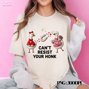 May include: Cream-colored t-shirt with a graphic featuring two cartoon geese in sweaters, a horseshoe magnet, and the text "CAN'T RESIST YOUR HONK." The geese are surrounded by hearts and stars.