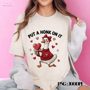 Puede incluir: Camiseta color crema con un dibujo de un ganso de dibujos animados con un suéter rojo y sosteniendo un objeto en forma de corazón. El texto "PUT A HONK ON IT" está encima del ganso. Corazones y estrellas rojas rodean al ganso, creando un tema de San Valentín.