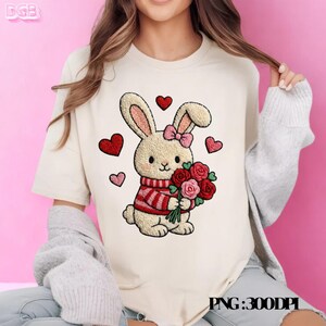 Peut inclure: T-shirt crème avec un lapin brodé tenant un bouquet de roses rouges. Le lapin porte un nœud rose et un pull rayé rouge et blanc. Des cœurs rouges et roses sont dispersés autour du motif du lapin.