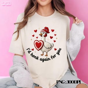 May include: A cream-coloured t-shirt with a cartoon goose wearing a red hat and holding a heart. The text "I'd honk again for you" is written around the goose. Red hearts and gold crosses surround the goose, creating a Valentine's Day theme.