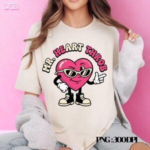 May include: Cream-colored t-shirt featuring a cartoon heart character with sunglasses and the words "MR. HEART THROB" in a circular design. The heart is pink with black accents, wearing sunglasses, and pointing with a white-gloved hand.