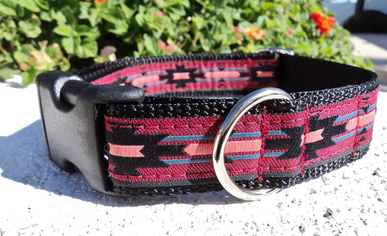 Pu&ograve; includere: Un collare per cani nero con un motivo geometrico rosa e blu. Il collare ha una fibbia in plastica nera e un anello a D in metallo argentato.