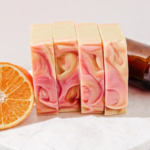 May include: Four bars of handmade soap with a swirl of pink and white. The soap is on a white marble surface with a half orange slice next to it.