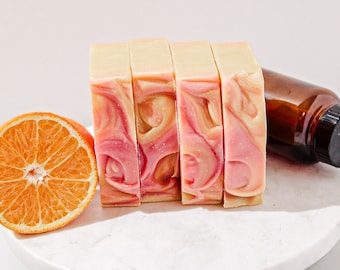 May include: Four bars of handmade soap with a swirl of pink and white. The soap is on a white marble surface with a half orange slice next to it.