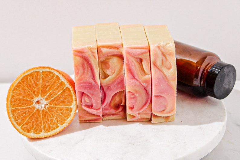 May include: Four bars of handmade soap with a swirl of pink and white. The soap is on a white marble surface with a half orange slice next to it.