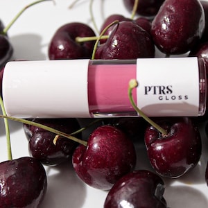 May include: A white tube of lip gloss with the text "PTRS GLOSS" sits among a pile of fresh, dark red cherries. The lip gloss contains a pink liquid. The cherries have green stems and are on a white surface.