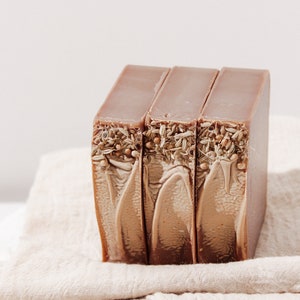 May include: Three bars of brown soap with dried herbs and seeds embedded in the top. The soap bars are sitting on a beige linen towel.
