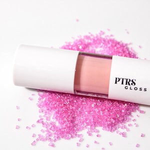 May include: A tube of PTRS Gloss lip gloss in a light pink shade, with a white cap and label, rests on a bed of bright pink sugar crystals. The label displays the brand name in black text.