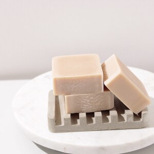 May include: Three bars of light brown soap stacked on a gray soap dish. The soap dish is on a white marble surface.