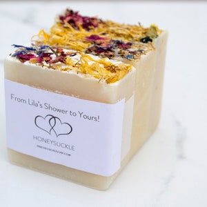 May include: Three bars of white soap with dried flowers and herbs embedded in the top. The soap is wrapped in a white label with the text "From Lila's Shower to Yours!" and "Honeysuckle".