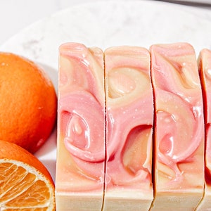 May include: Four bars of handmade soap with a swirl design in shades of pink and orange. The soap is arranged in a row on a white surface.