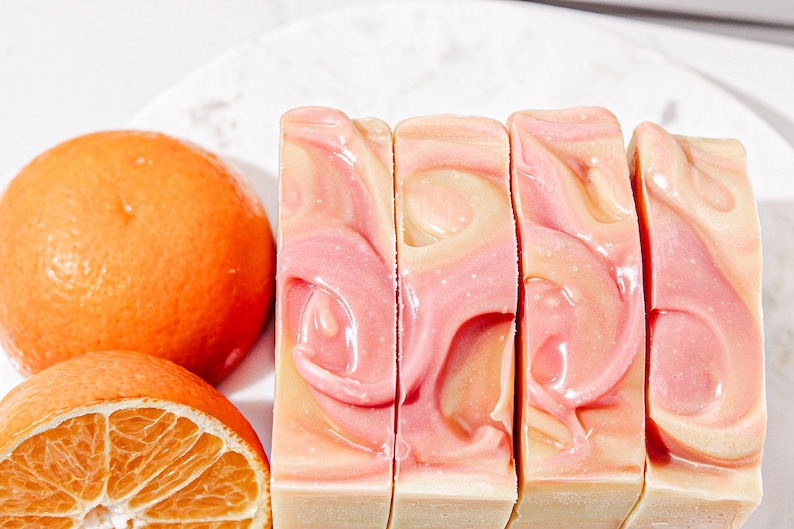 May include: Four bars of handmade soap with a swirl design in shades of pink and orange. The soap is arranged in a row on a white surface.