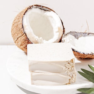 May include: Two bars of white soap with oatmeal on a white marble surface. The soap is next to two halves of a coconut shell.