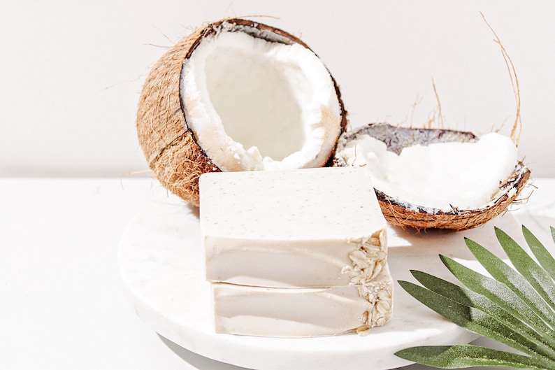 May include: Two bars of white soap with oatmeal on a white marble surface. The soap is next to two halves of a coconut shell.