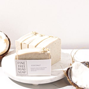 May include: A stack of white and beige handmade soap bars with oatmeal on top. The soap is labelled "Pine Tree Road Soap" and "Coconut". The soap is sitting on a white marble surface with a half coconut shell next to it.