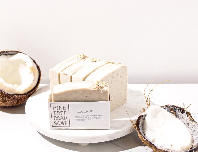 May include: A stack of white and beige handmade soap bars with oatmeal on top. The soap is labelled "Pine Tree Road Soap" and "Coconut". The soap is sitting on a white marble surface with a half coconut shell next to it.