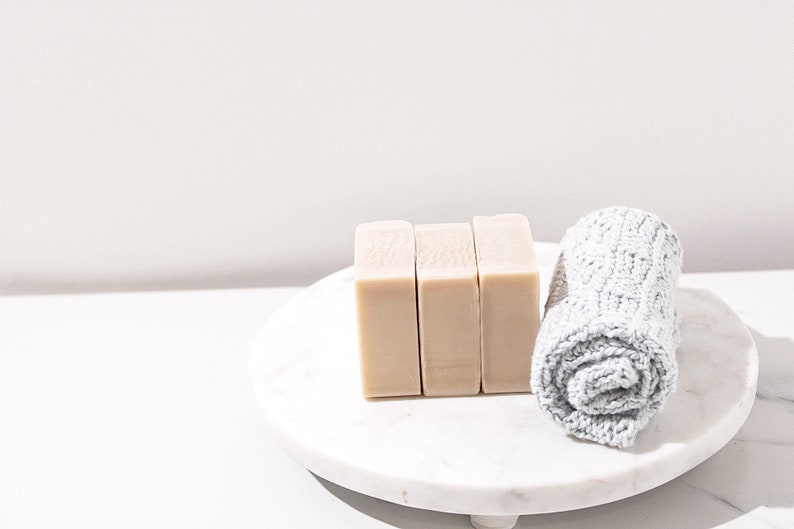 May include: Three bars of beige soap on a white marble pedestal with a rolled up gray hand towel.