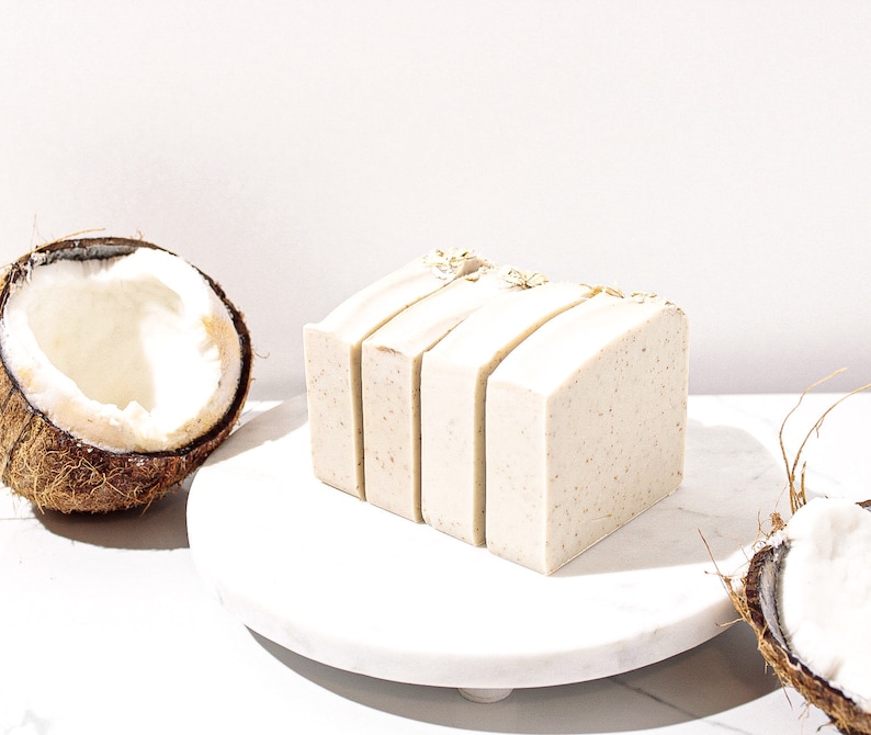 May include: Four bars of natural soap with a light brown colour and a grainy texture. The soap is arranged in a stack on a white marble surface. A half coconut shell is visible in the background.
