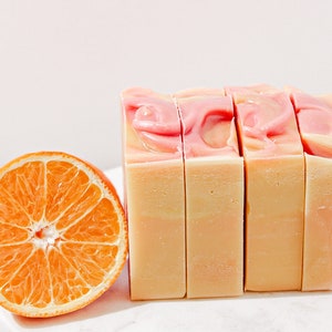 May include: Four bars of handmade soap with a pink and white swirl design. The soap is sitting on a white surface next to a slice of orange.
