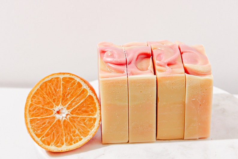 May include: Four bars of handmade soap with a pink and white swirl design. The soap is sitting on a white surface next to a slice of orange.