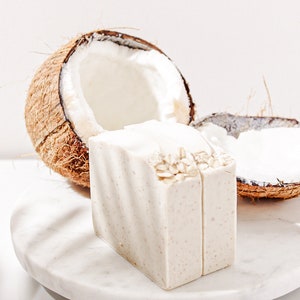 May include: Two bars of white soap with oatmeal on top, sitting on a white marble surface next to a halved coconut shell.