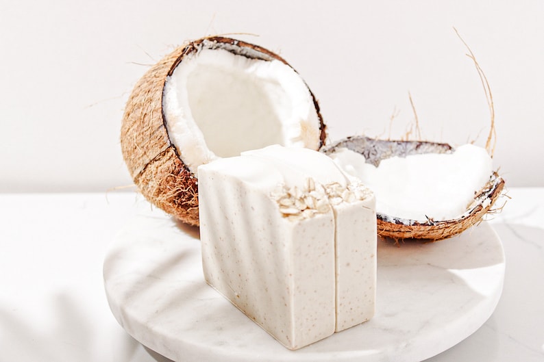 May include: Two bars of white soap with oatmeal on top, sitting on a white marble surface next to a halved coconut shell.