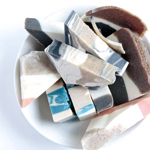 May include: A white ceramic dish filled with various handmade soap bars. The soap bars are in different colors and patterns, including brown, white, black, and blue.